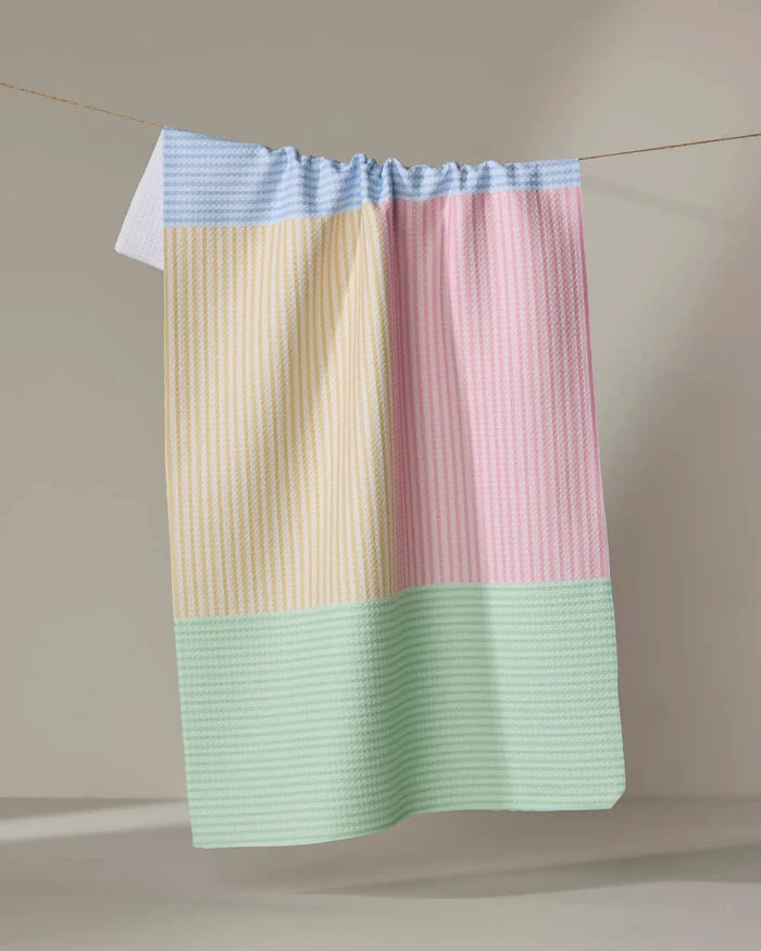 The Modern Dad’s Mr. Fun Stripes kitchen tea towel features a pastel patchwork in blue, yellow, pink, and green. Ultra-absorbent and stylish, it hangs on a line against a plain beige background—practical for daily use.