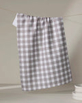 A chocolate and white checkered recycled kitchen towel by The Buy Guide (Multi Gingham - Chocolate) hangs on a clothesline, with a stack of white plates partially visible in the lower right corner against a neutral background.