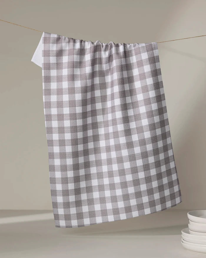 A chocolate and white checkered recycled kitchen towel by The Buy Guide (Multi Gingham - Chocolate) hangs on a clothesline, with a stack of white plates partially visible in the lower right corner against a neutral background.