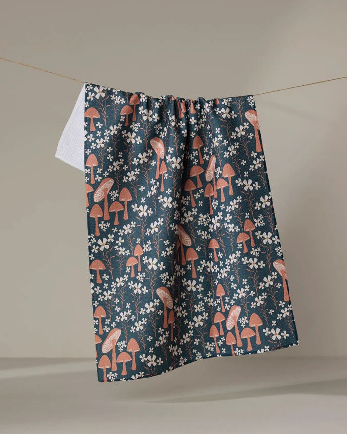 The Mushroom Grove tea towel by Charlie Rowan Designs, made from recycled materials, showcases an orange mushroom and white flower pattern as it hangs on a clothesline against a neutral backdrop.
