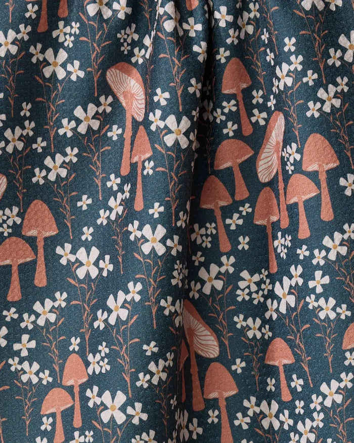 The Mushroom Grove kitchen towel by Charlie Rowan Designs features a dark background with light brown mushrooms and small white flowers with green stems, offering a whimsical, nature-inspired touch to your kitchen.
