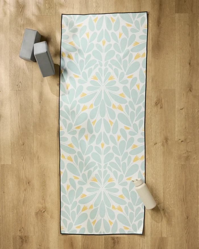 The Rebecca Woolbright Mustard Pinwheel yoga mat with a floral design is laid out on a wooden floor, topped with a non-slip towel. Two gray yoga blocks and a white water bottle are placed nearby.