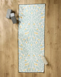 The Rebecca Woolbright Mustard Pinwheel yoga mat with a floral design is laid out on a wooden floor, topped with a non-slip towel. Two gray yoga blocks and a white water bottle are placed nearby.