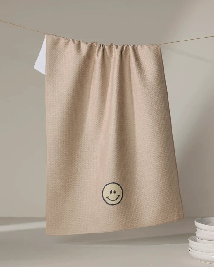The "My Happy Place" kitchen towel by Kelsi Fullmer, featuring a beige textured design and an embroidered black smiley face, hangs on a clothesline above stacked white dishes, adding charm to your eco-friendly kitchen.