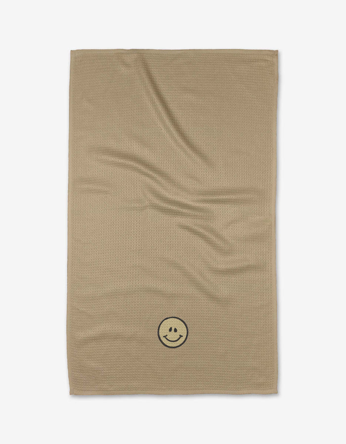 The My Happy Place tea towel by Kelsi Fullmer is beige, rectangular, made from absorbent recycled materials, and features a subtle texture and a black smiley face in one corner, shown on a white background.