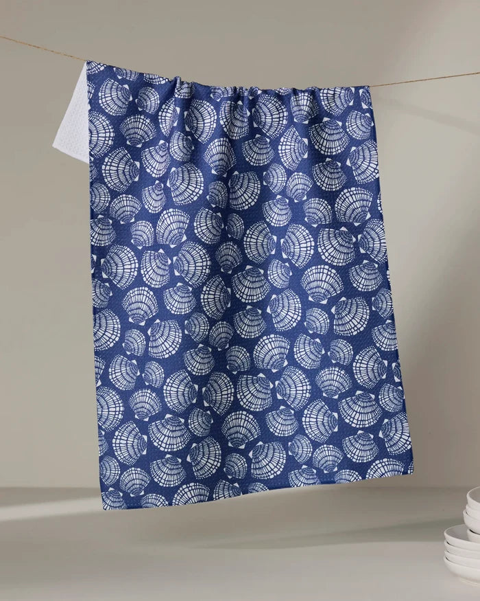 The Kris Ruff Nautical Seashells recycled kitchen towel, featuring a blue and white seashell design, hangs on a clothesline against a neutral background with white dishes stacked in the lower right corner.