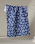 The Kris Ruff Nautical Seashells recycled kitchen towel, featuring a blue and white seashell design, hangs on a clothesline against a neutral background with white dishes stacked in the lower right corner.