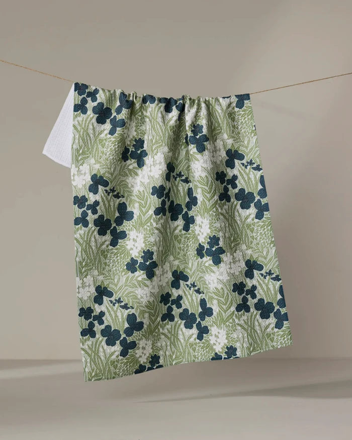 The Katie O'Shea Navy And Sage Meadow towel, featuring a green and white floral pattern with dark blue flowers, hangs on a clothesline against a plain beige background.