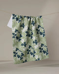 The Katie O'Shea Navy And Sage Meadow towel, featuring a green and white floral pattern with dark blue flowers, hangs on a clothesline against a plain beige background.
