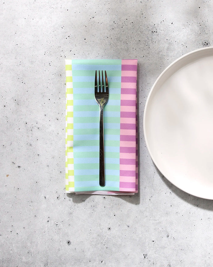 A fork rests on GEOMETRY's Neon Nights pastel striped recycled fabric napkin, set beside a plain white plate on a light gray speckled surface.