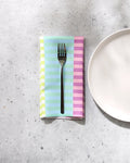 A fork rests on GEOMETRY's Neon Nights pastel striped recycled fabric napkin, set beside a plain white plate on a light gray speckled surface.