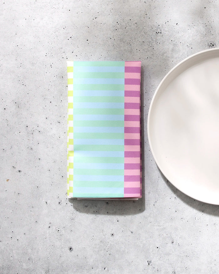 A folded GEOMETRY Neon Nights pastel-striped napkin in green, blue, and pink rests on a light gray speckled surface next to a white plate.