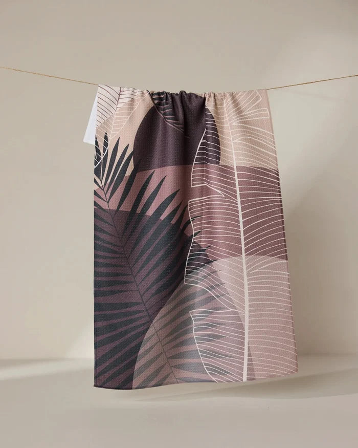 The GEOMETRY Neutral Hills tea towel, featuring a pink, mauve, and beige botanical leaf design, dries quickly and hangs on a string against a light beige background.