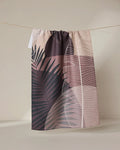 The GEOMETRY Neutral Hills tea towel, featuring a pink, mauve, and beige botanical leaf design, dries quickly and hangs on a string against a light beige background.