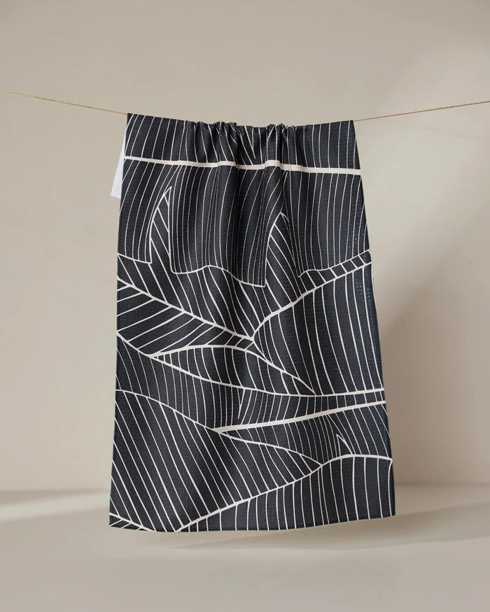 The GEOMETRY Neutral Palms tea towel, featuring a white geometric leaf pattern on black, hangs from a string against a plain beige background.