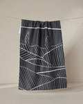 The GEOMETRY Neutral Palms tea towel, featuring a white geometric leaf pattern on black, hangs from a string against a plain beige background.