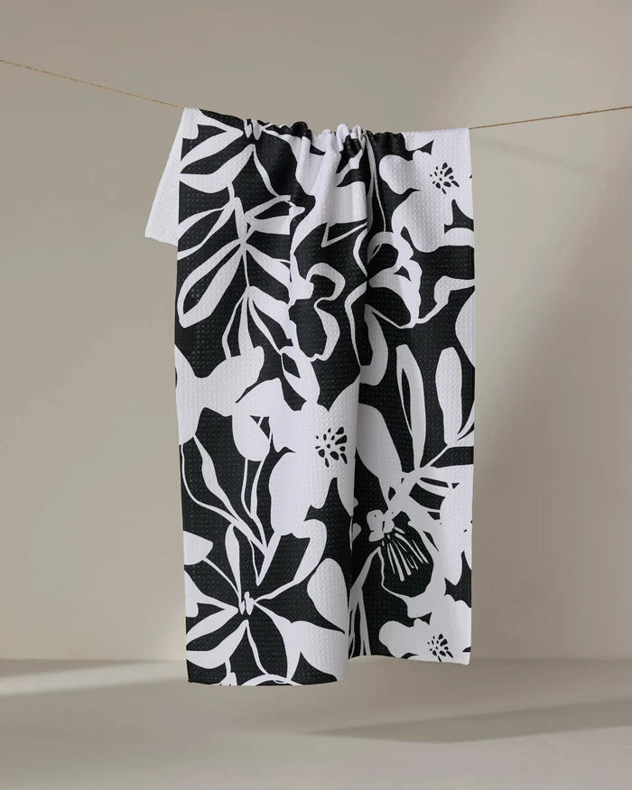 The Night Meadow tea towel by Marina Solodka, featuring a bold black and white abstract floral design and made from recycled materials, hangs on a string against a neutral background.