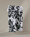The Night Meadow tea towel by Marina Solodka, featuring a bold black and white abstract floral design and made from recycled materials, hangs on a string against a neutral background.