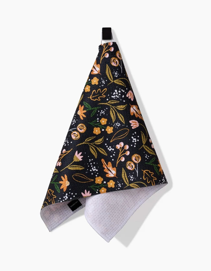 The Megan Roy Nighttime Botanical bar towel features a colorful floral pattern in pink, yellow, and green on black, hangs from a black loop, and is made of Geoweave fabric with a white textured underside for absorbency and durability.