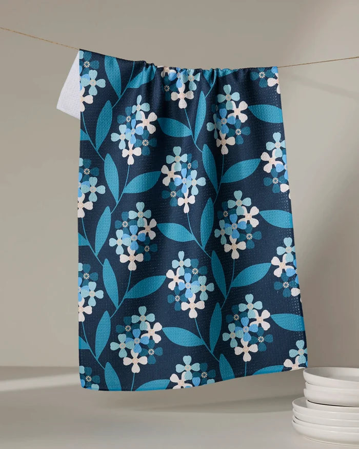 The Nighttime Florals tea towel by Blissful Flamingo, featuring a blue and white floral pattern, hangs on a string. Soft lighting highlights stacked white plates, adding an elegant touch to your kitchen décor.