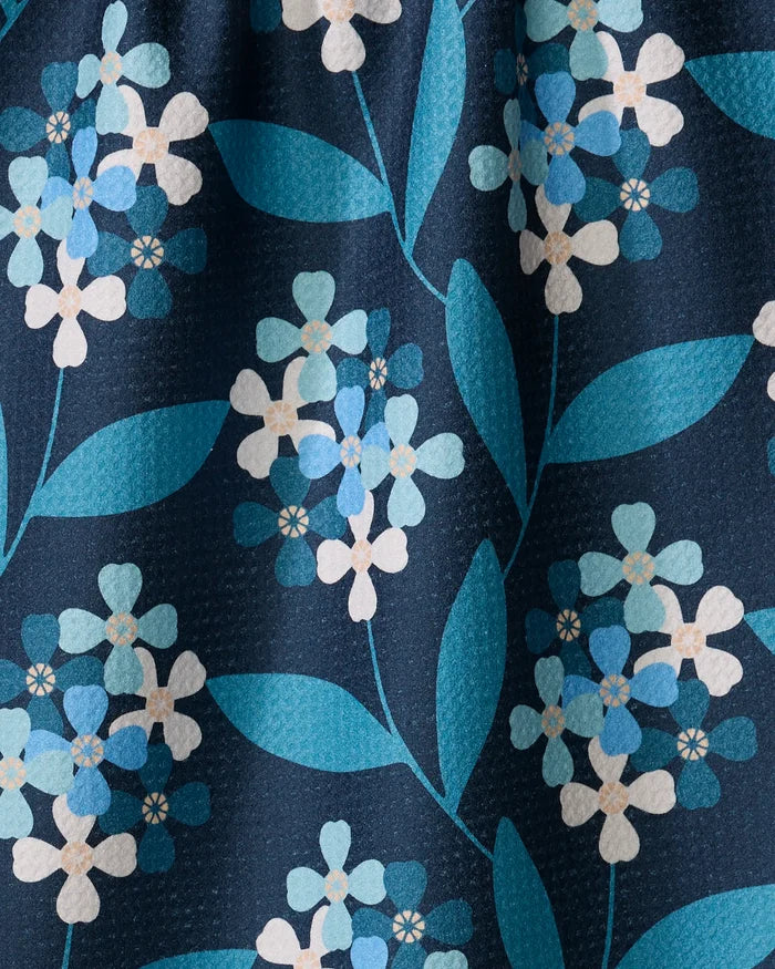 The Blissful Flamingo "Nighttime Florals" towel showcases a close-up floral pattern with blue and white flowers, green leaves on dark blue. Ultra-absorbent, it's made from recycled materials with a subtly textured surface.