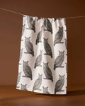 The Geometry Nocturne tea towel, featuring a repeating black owl print on white, hangs against a plain brown background.