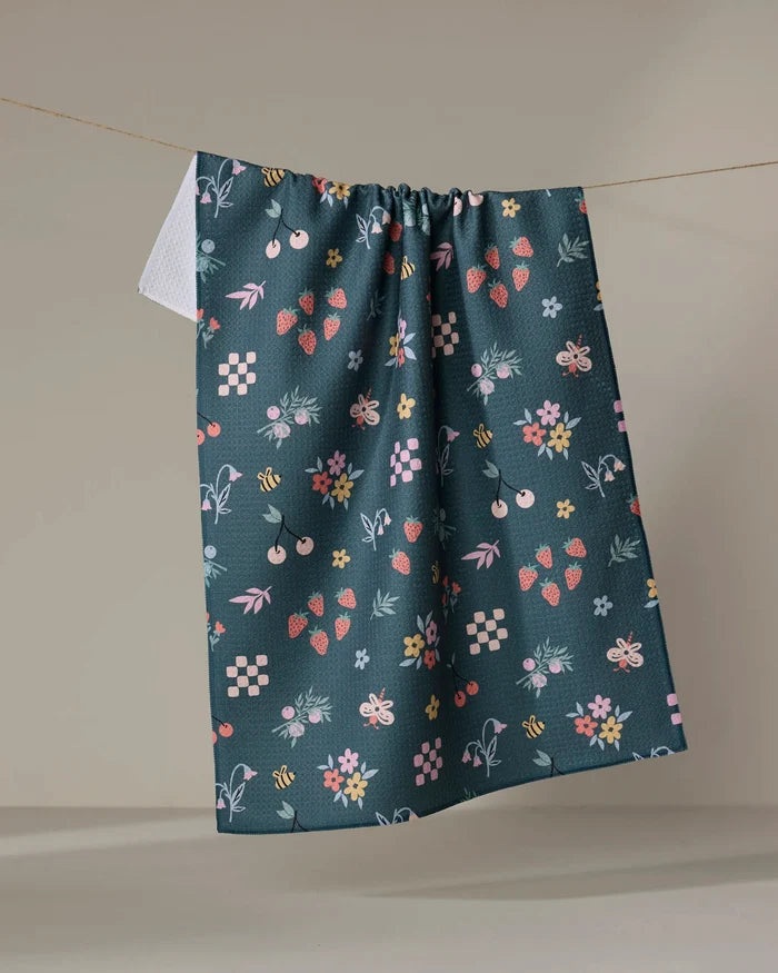 The North Meadow tea towel by Charlie Rowan Designs is a dark green, recycled towel with vibrant strawberries, bees, flowers, cherries, and leaves, displayed hanging against a neutral backdrop.