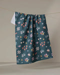 The North Meadow tea towel by Charlie Rowan Designs is a dark green, recycled towel with vibrant strawberries, bees, flowers, cherries, and leaves, displayed hanging against a neutral backdrop.