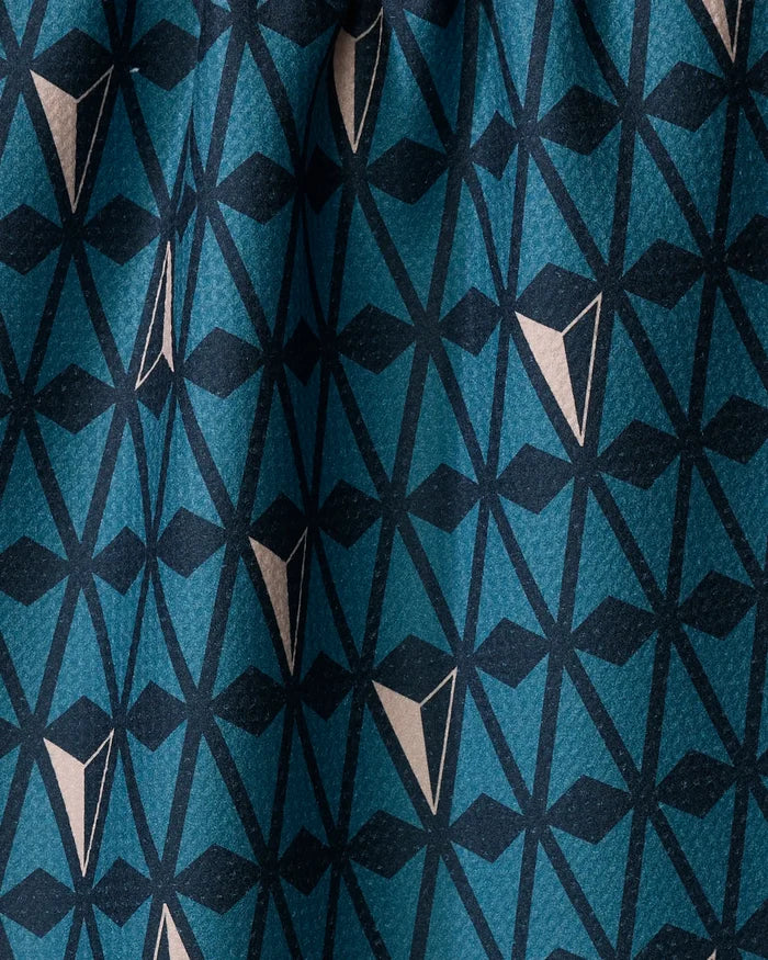 A close-up of Twig + Spark's Northbound tea towel shows its quick-drying fabric with a dynamic geometric pattern in teal, navy, and beige triangles and lines. The repeating design appears slightly folded, perfect for modern kitchens.