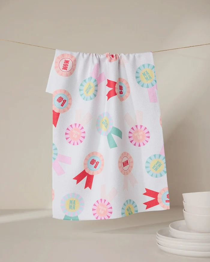 The “Number One Mom” tea towel by GEOMETRY, made from recycled materials, features colorful ribbon designs and “MOM” and “MAMA” text. It hangs above white dishes, offering an eco-friendly accent to a neutral kitchen.