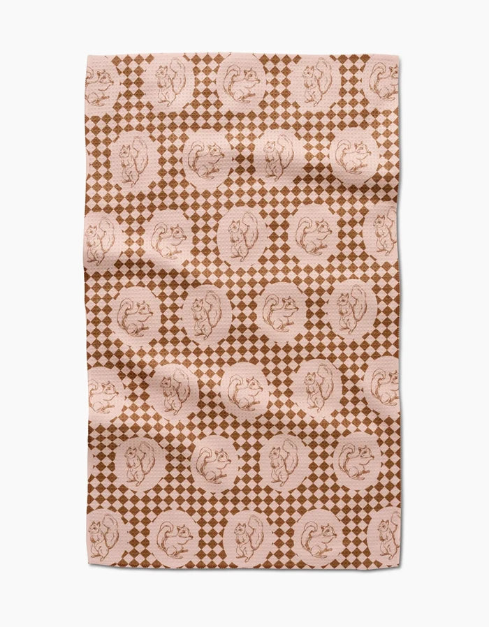 The “Nuts About You” tea towel by Aubrey Fairchild features a brown and beige circular squirrel design over small diamonds. Made of quick-drying fabric, it appears slightly wrinkled and is displayed on a white surface.