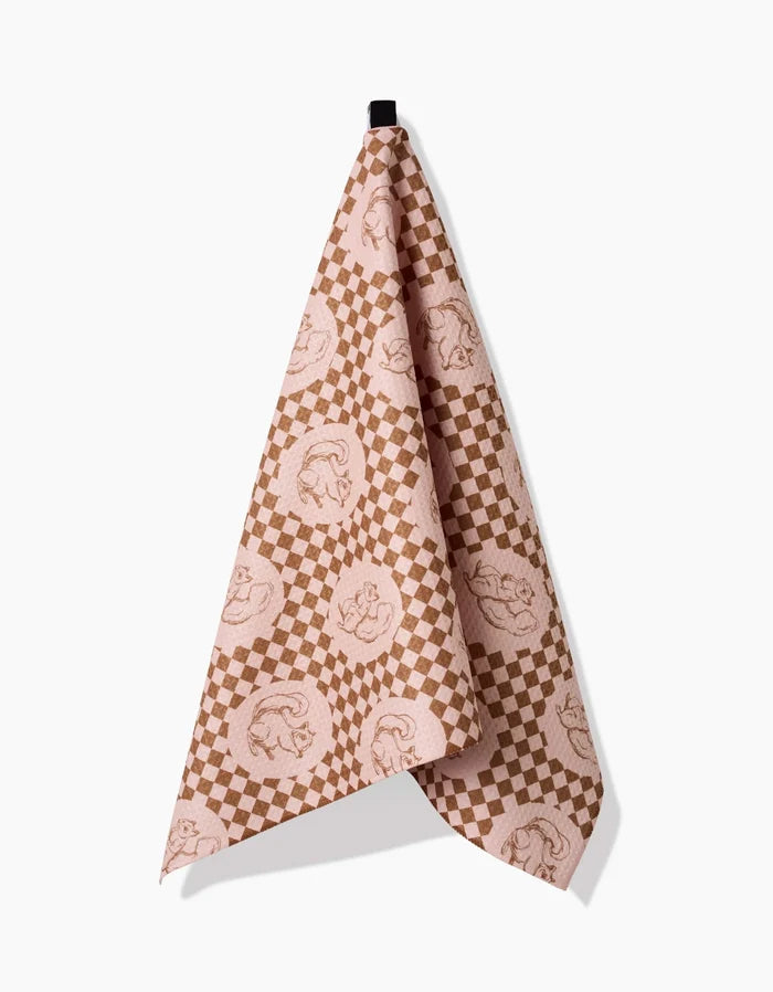 The Aubrey Fairchild Nuts About You tea towel, featuring pink and brown checkered circles and made from recycled materials, hangs on a black hook against a plain white background.
