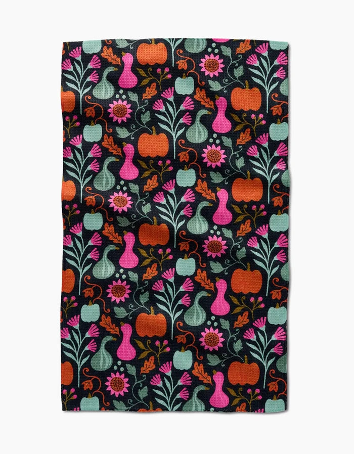 The Oakley kitchen towel by Amy MacCready features a vibrant folk art design of pumpkins, gourds, flowers, and leaves in orange, pink, teal, and green on a black background for a whimsical touch.