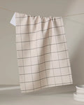 The Oat Grid tea towel by Kelsi Fullmer, featuring a beige base with a simple black grid design, hangs on a string with its white corner folded over. Stacked white plates sit in the lower right against a neutral backdrop.
