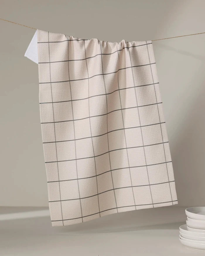 The Oat Grid tea towel by Kelsi Fullmer, featuring a beige base with a simple black grid design, hangs on a string with its white corner folded over. Stacked white plates sit in the lower right against a neutral backdrop.