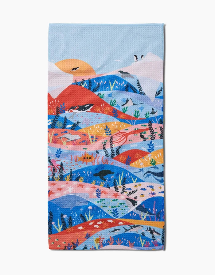 The Oceans bar towel by Ceyda Alasar is made from recycled materials and showcases a vibrant ocean scene with waves, marine life, plants, and a sunset in blue, orange, and red tones.