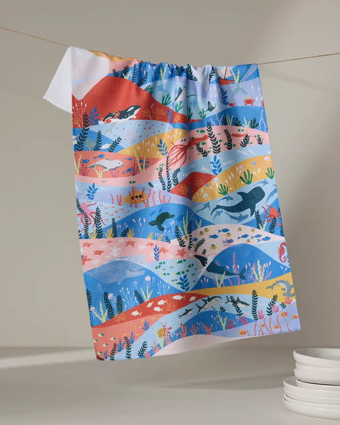 A vibrant recycled kitchen towel from Ceyda Alasar's "Oceans" collection, featuring ocean-themed patterns in blues, reds, yellows, and oranges, hangs on a line above stacked white plates.