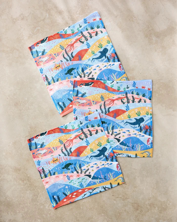 Three colorful "Oceans" kitchen towels by Ceyda Alasar, featuring a repeating dinosaur and plant pattern in blue, orange, yellow, and red tones, are displayed on a light stone surface.
