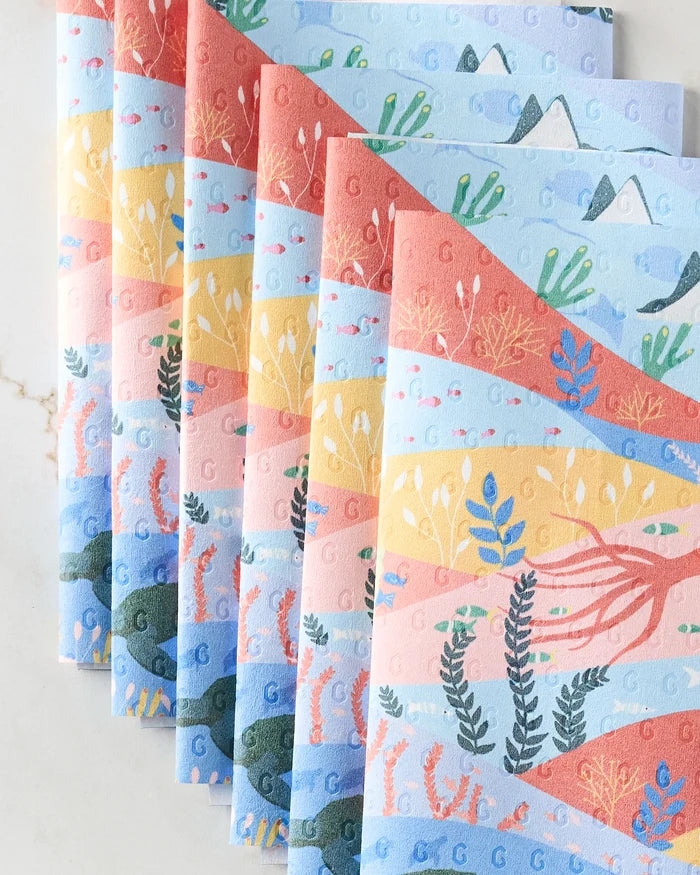Oceans by Ceyda Alasar: eco-friendly paper towels in soft pastel blue, pink, and orange with illustrated underwater and desert scenes—fish, sea plants, coral, mountains, and cacti—artfully arranged in a fan pattern.