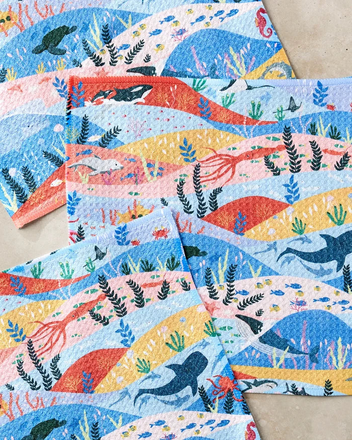 The "Oceans" dishcloths by Ceyda Alasar feature recycled materials and vibrant ocean scenes with whales, turtles, fish, seaweed, and coral on wavy blue, yellow, and pink backgrounds over a light base.