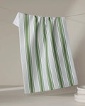 Aubrey Fairchild’s "Of the Same Stripe" tea towel, crafted from recycled materials, hangs on a clothesline above stacked white plates in the lower right corner. Its white fabric features vertical green and thin black stripes against a neutral backdrop.