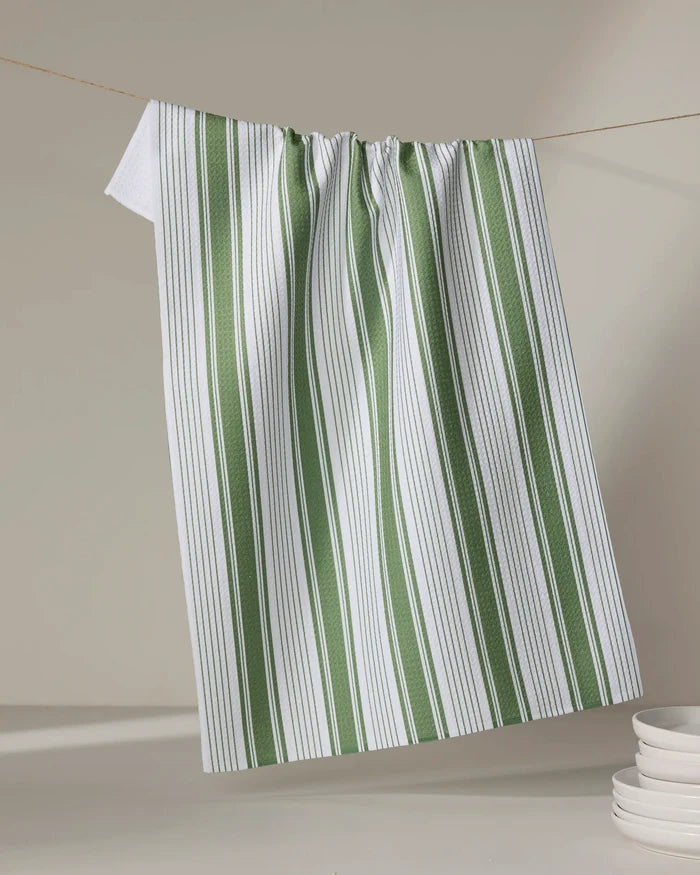 Aubrey Fairchild’s "Of the Same Stripe" tea towel, crafted from recycled materials, hangs on a clothesline above stacked white plates in the lower right corner. Its white fabric features vertical green and thin black stripes against a neutral backdrop.