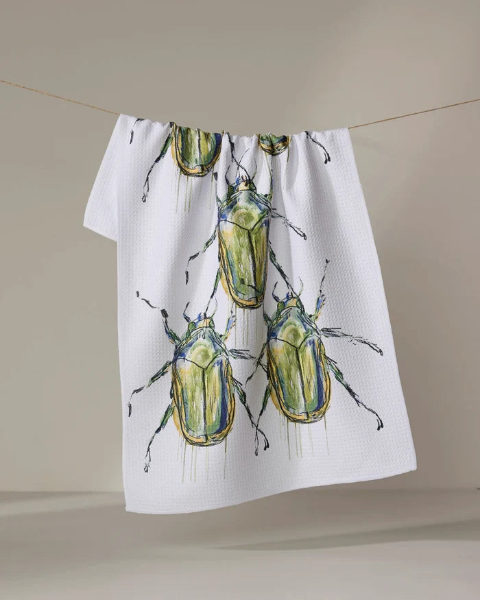 The Olive Beetles kitchen towel by Taryn Brennan features eco-friendly white fabric with large green and yellow watercolor beetles, shown hanging on a clothesline against a plain background.