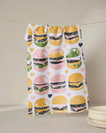 The Geometry On The Grill tea towel, featuring a vibrant hamburger and heart pattern, hangs above stacked white plates against a neutral backdrop.