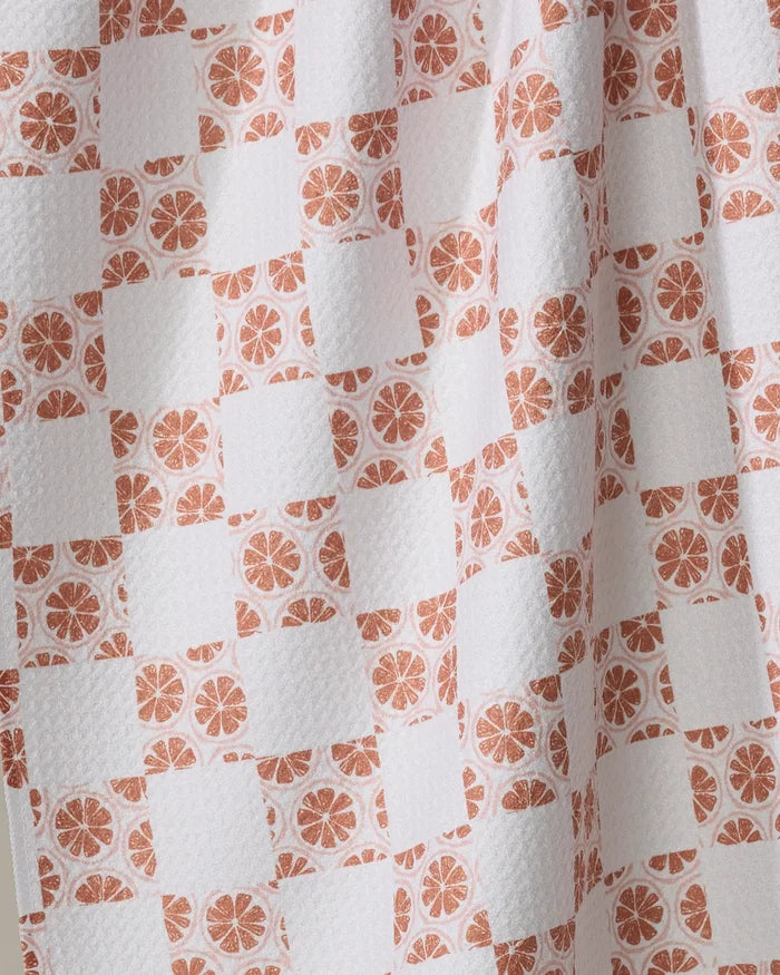 A close-up of Orange Checker by Teri the Butter shows textured white fabric made from recycled materials, with a checkered pattern of plain white squares and orange citrus slices. Soft draping creates gentle folds and shadows—ideal for a tea towel.