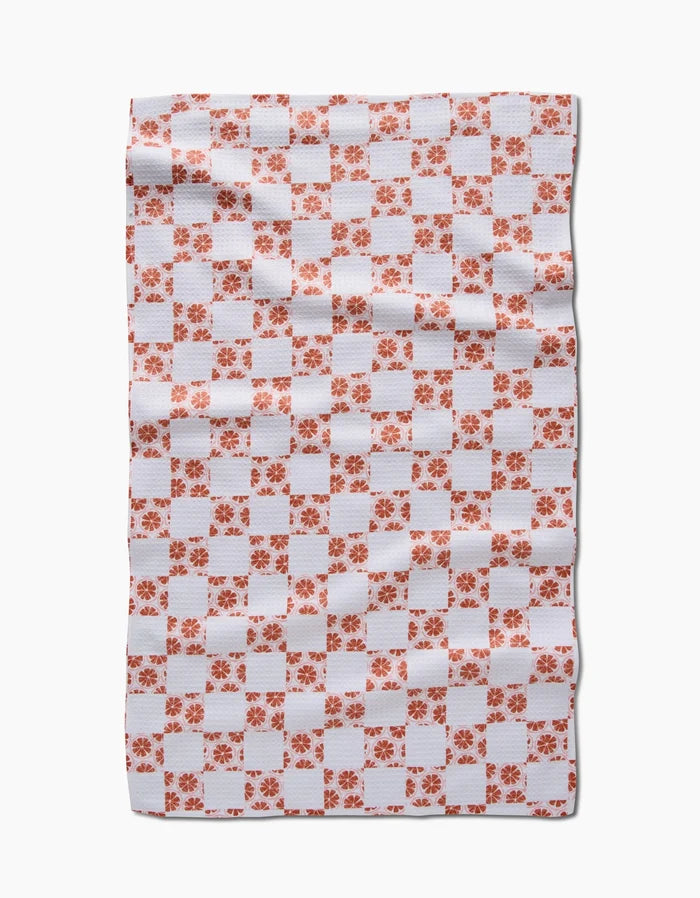 The Orange Checker by Teri the Butter is a rectangular white tea towel with an orange checkered pattern and floral designs, crafted from Geoweave™ fabric. Cloth appears slightly wrinkled, shown on a plain white background.