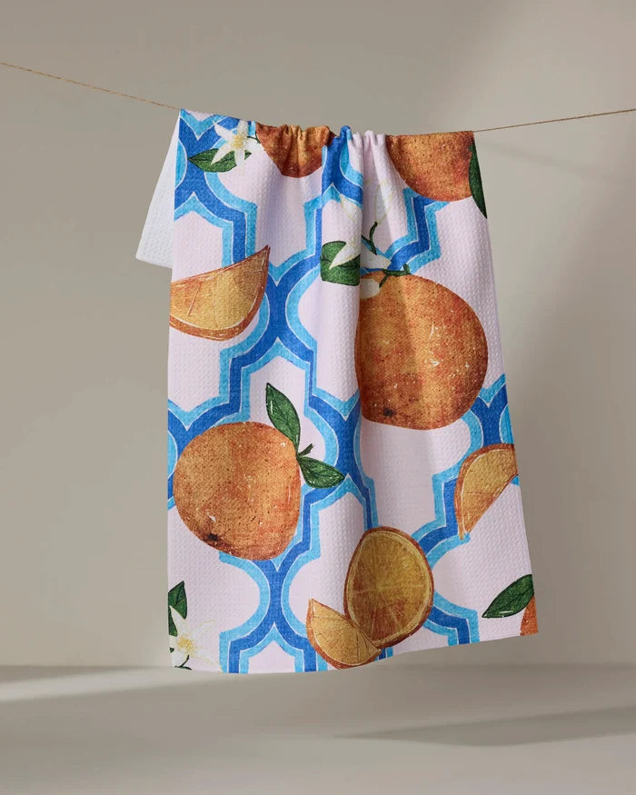 The Pascale Dilger "Orange Trellis" kitchen tea towel features whole and sliced oranges with green leaves on a blue and white geometric background, adding an absorbent and stylish touch to your home.