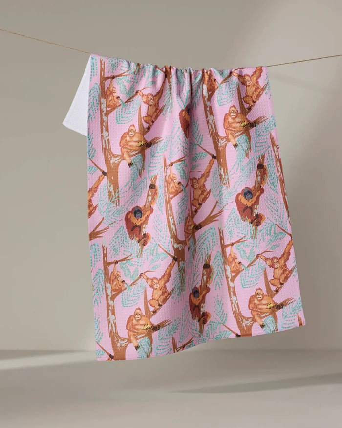 The "Orangutan Clan" kitchen towel by Elisabeth Wing features playful orangutans hanging from branches, is made from recycled materials, and is shown draped over a clothesline against a neutral background.