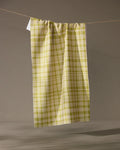 The Orchard Plaid Green tea towel by Geometry hangs vertically from a string against a gray background. Made from recycled materials, its lightweight fabric is gathered at the top for an eco-friendly, stylish touch.