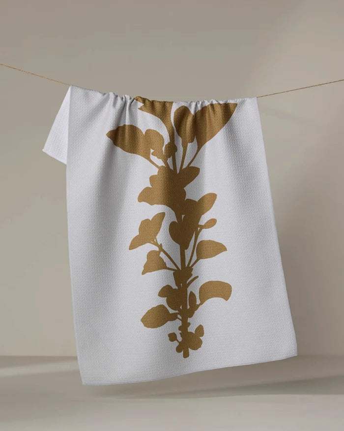 PrintedPlant’s “Oregano” is a white textured tea towel featuring a bold gold botanical silhouette, made from recycled materials and displayed hanging on a thin string against a neutral background.